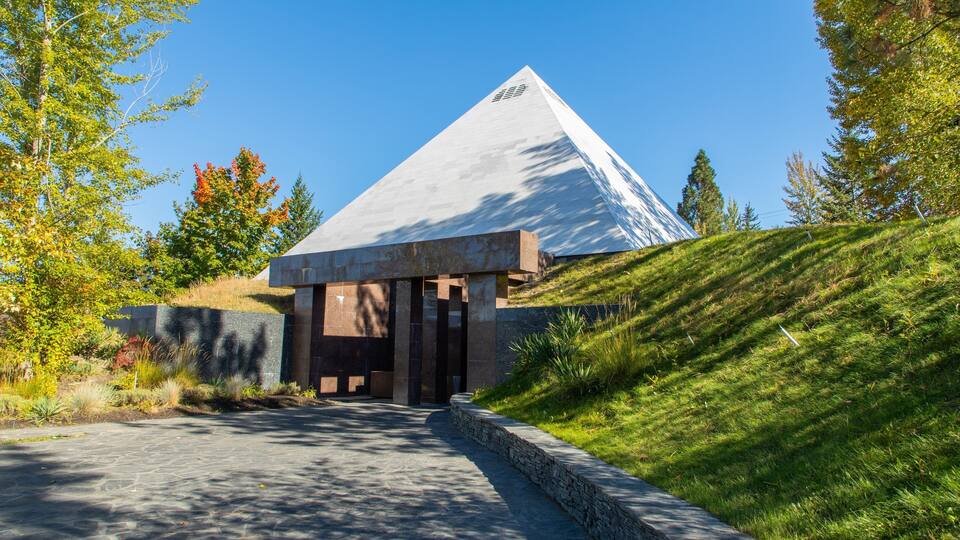 A modern building with a pyramid-like roof, surrounded by a landscaped area with trees and a sloped hill.