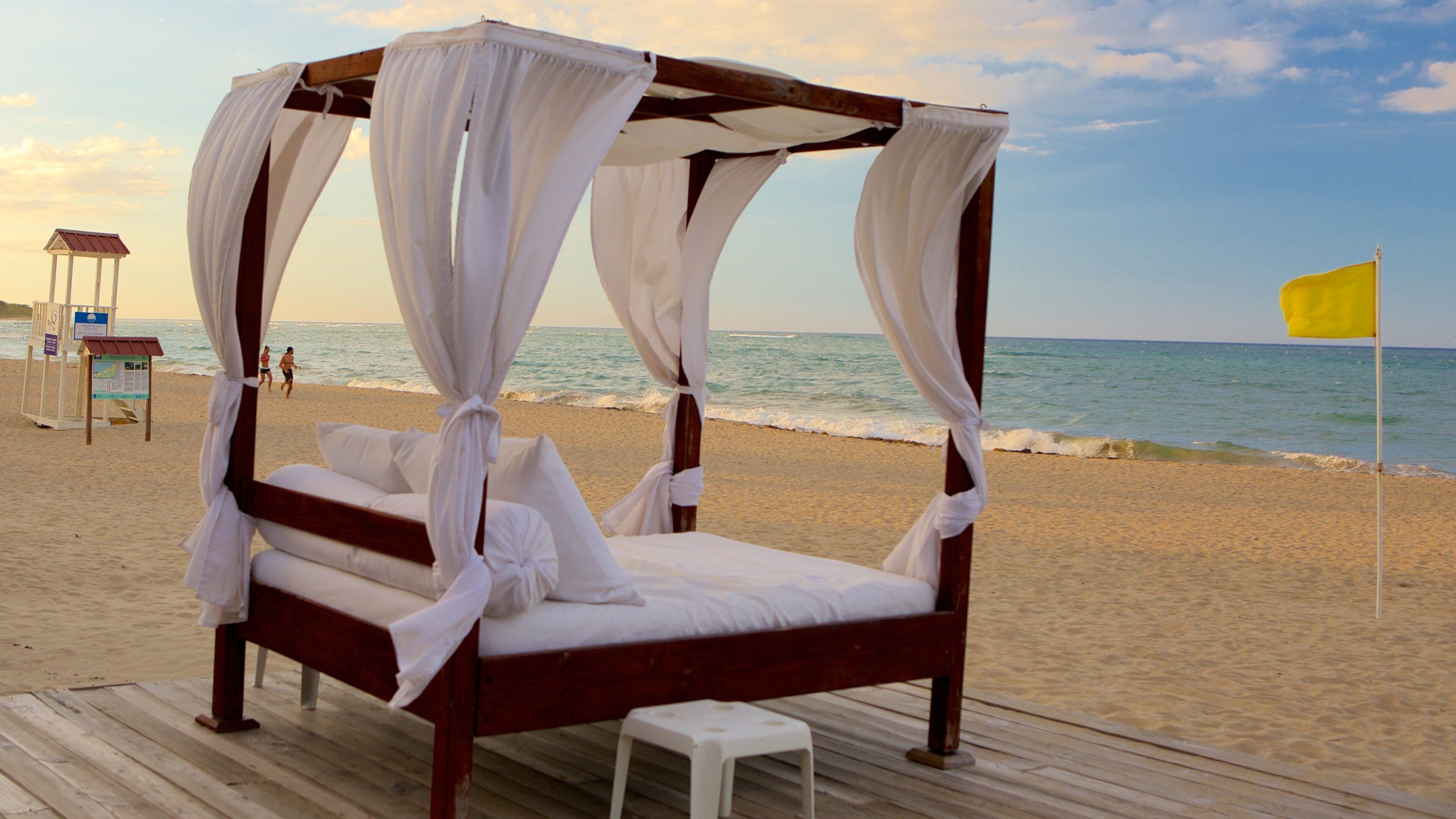 A wooden four-poster bed with white curtains on a wooden deck at the beach.