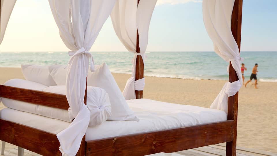 A wooden bed with white bedding and canopy, set on a wooden deck by a beach.