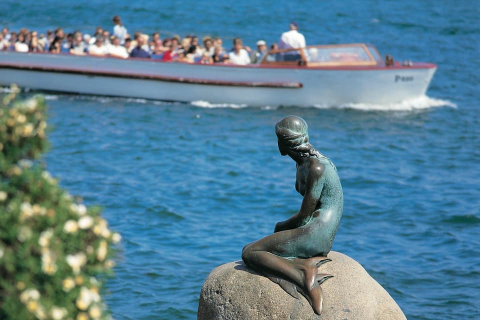A bronze statue of a seated figure on a rock by the water, with a boat carrying passengers in the background.