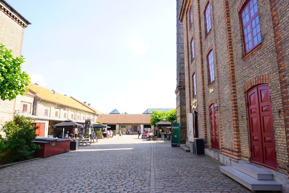 Een geplaveid straatje in een historische stad met bakstenen gebouwen, terrasjes en een heldere lucht.