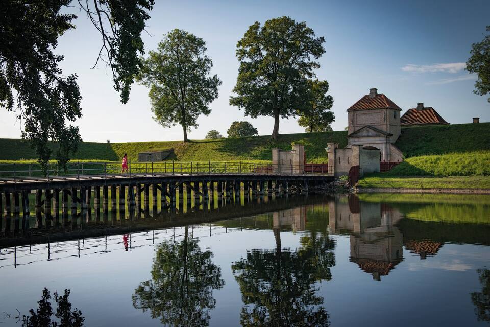 Un riu tranquil amb un pont de fusta, un petit edifici de pedra i arbres verds i frondosos.