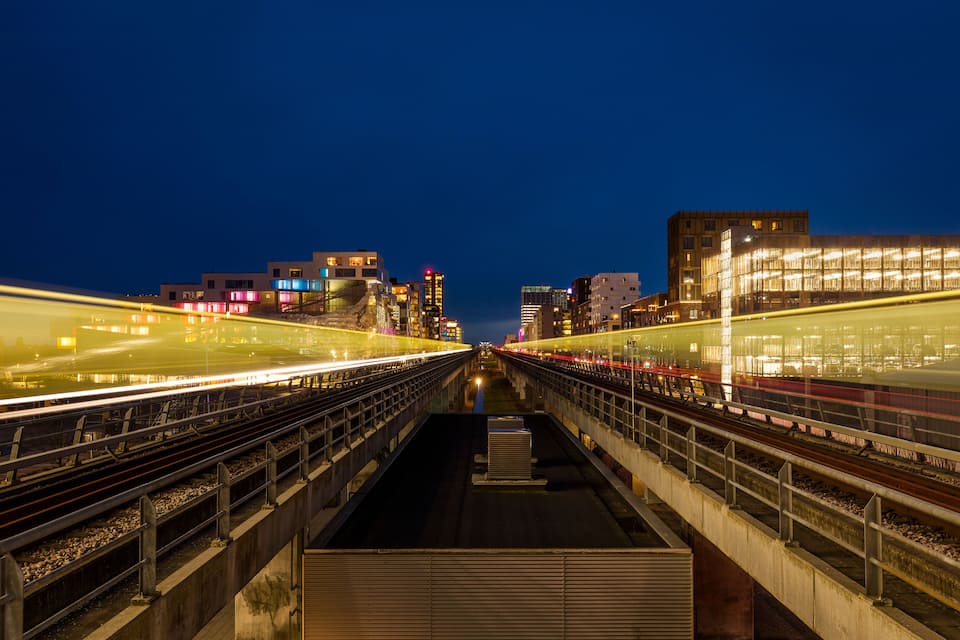 Ein Nachtbild eines Bahnhofs mit mehreren Gleisen und einer Stadtlandschaft im Hintergrund.