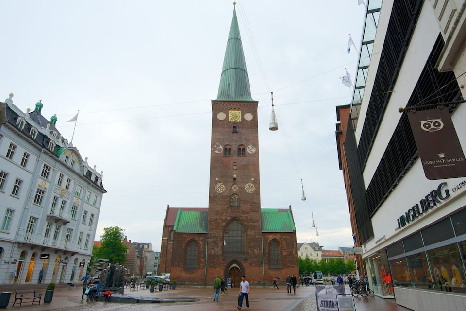Une église historique, dotée d’un flèche imposante et d’une tour horloge, entourée de bâtiments modernes et d’une place pavée.