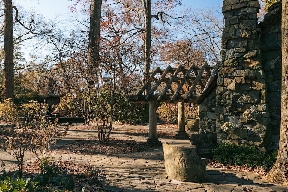 A stone structure with a wooden pergola, surrounded by trees and a stone bench.