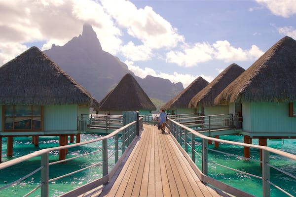 A wooden walkway between overwater bungalows with thatched roofs, leading to a mountain in the background.