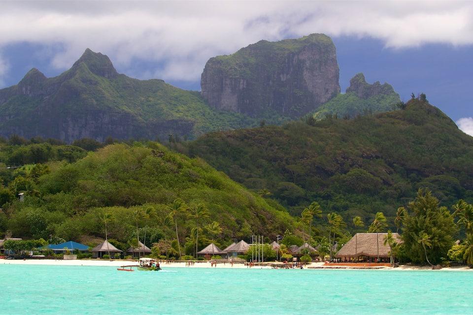 A tropical beach with clear turquoise waters, palm trees, and thatched-roof huts. In the background, there are lush green mountains.