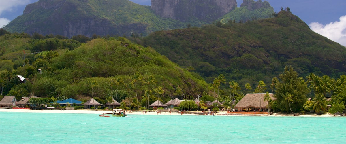 A tropical beach with clear turquoise waters, palm trees, and thatched-roof huts. In the background, there are lush green mountains.