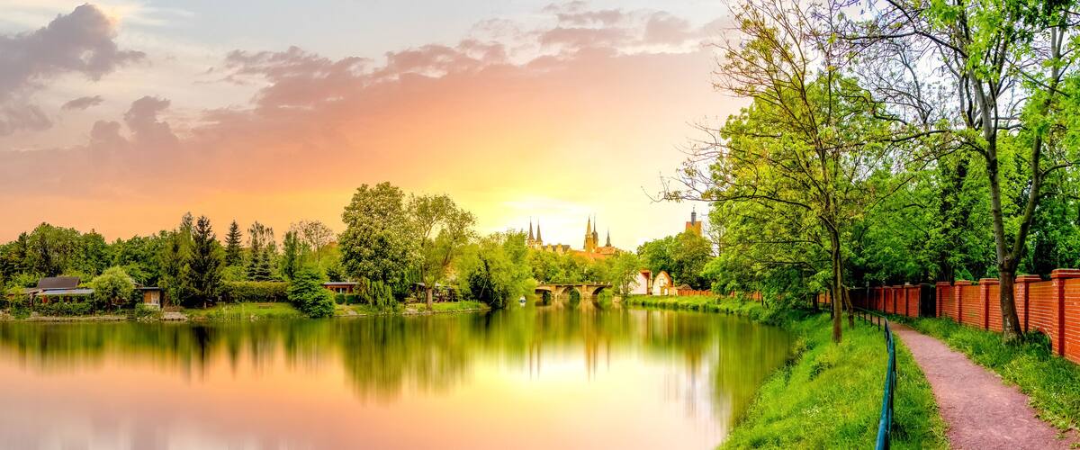 A calm river with a bridge, lush greenery, and a historic building in the background.