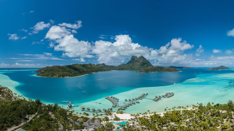 A tropical island with clear turquoise waters, a prominent mountain peak, and overwater bungalows.