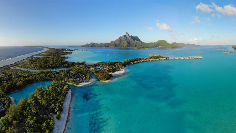 A tropical island with clear turquoise waters, a prominent mountain peak, and a resort area with overwater bungalows.