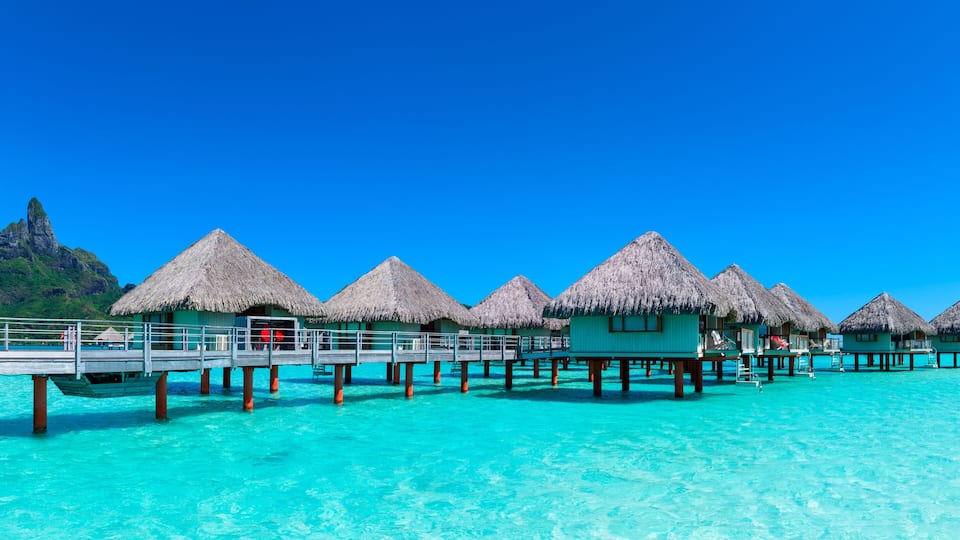 Overwater bungalows with thatched roofs, a wooden pier, and clear turquoise water.