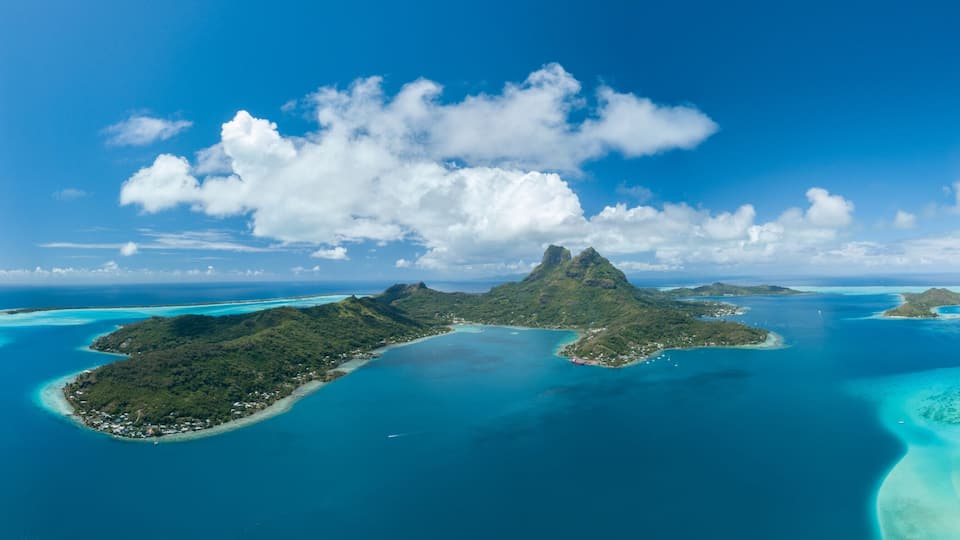 A tropical island landscape with clear turquoise waters, a prominent mountain peak, and lush greenery.
