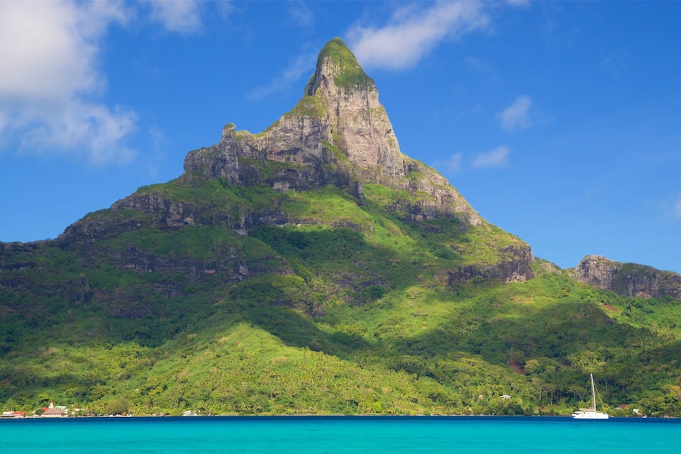 A prominent mountain peak with lush green vegetation, a clear blue sky, and a calm turquoise sea.