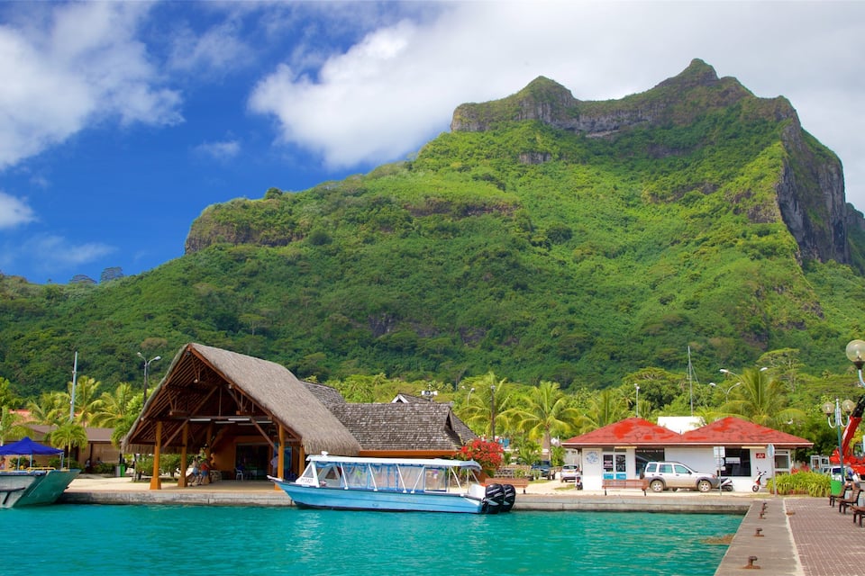 A tropical beach with a mountain in the background, a boat docked at the pier, and a small building with a thatched roof.