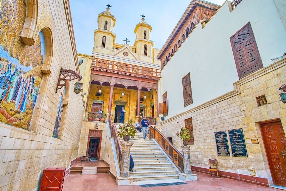A historic building with a staircase leading to a balcony, surrounded by ornate architecture and a mosaic wall.