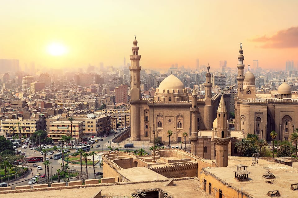 A cityscape with a prominent mosque featuring multiple domes and minarets, surrounded by dense urban buildings and palm trees.