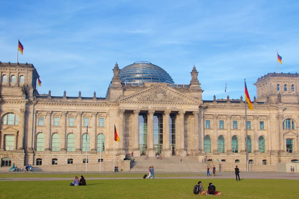 Un grand bâtiment historique, doté d’un dôme et de colonnes imposants, avec des drapeaux et des personnes au premier plan.