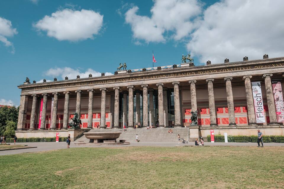 Un edificio clásico con columnas y estatuas, una bandera y gente caminando.