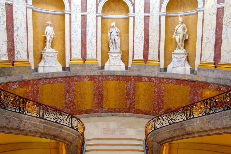 Una imponente escalera de mármol con detalles dorados que conduce a un balcón con estatuas.