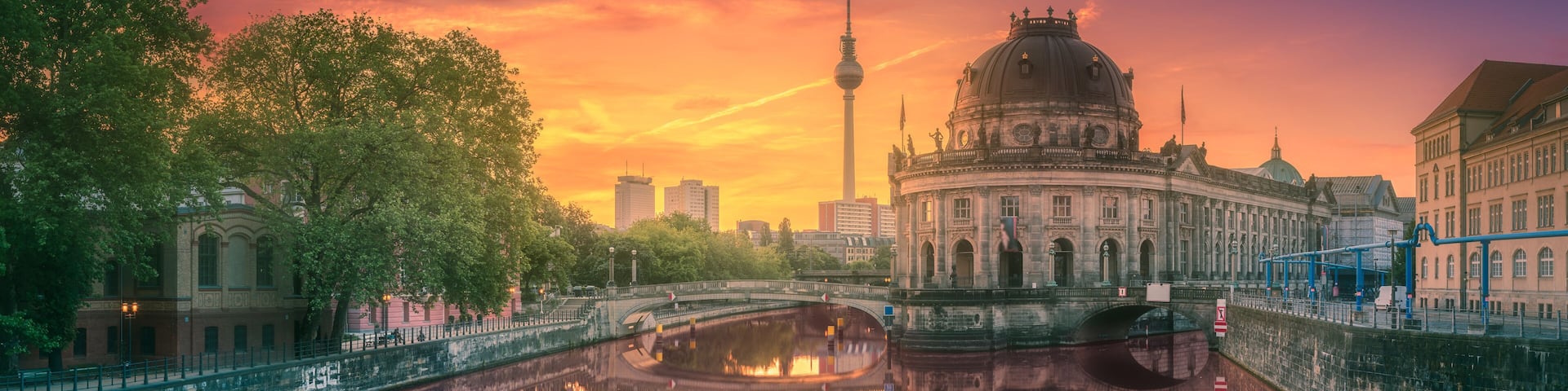 A river reflecting a historic building and a tower, surrounded by trees and urban structures.