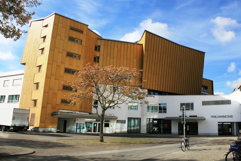 A modern building with a mix of brick and white facades, a tree in the foreground, and a clear blue sky.