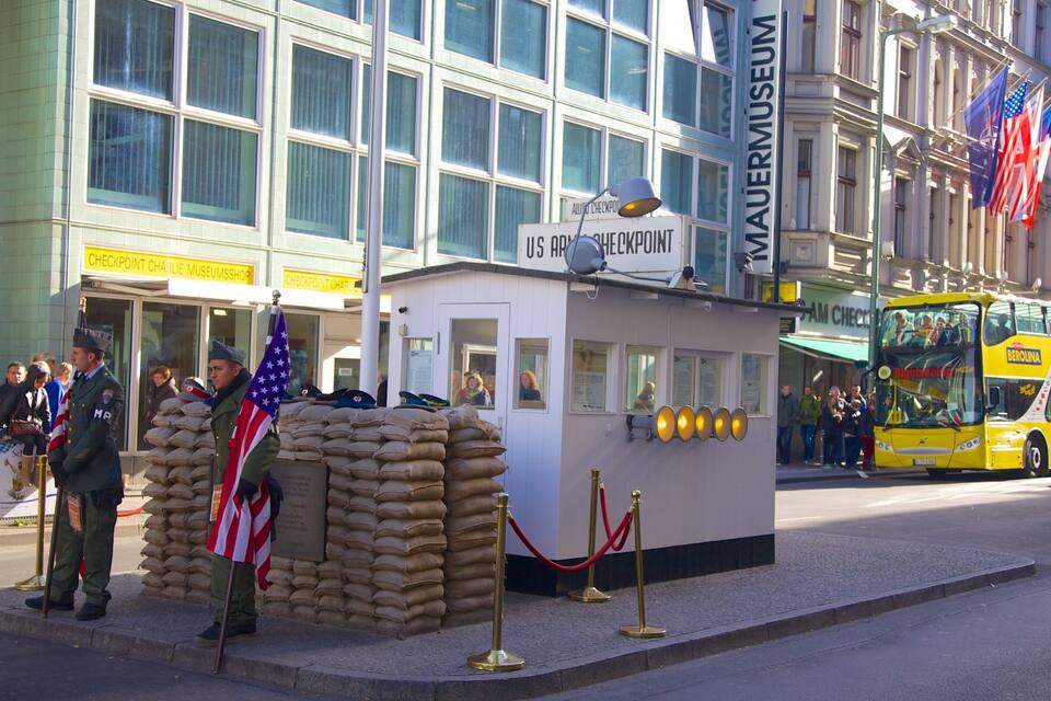 Een klein gebouw met de tekst 'MAUERMUSEUM', een bord van het Amerikaanse leger, een vlag en een bus op de achtergrond.