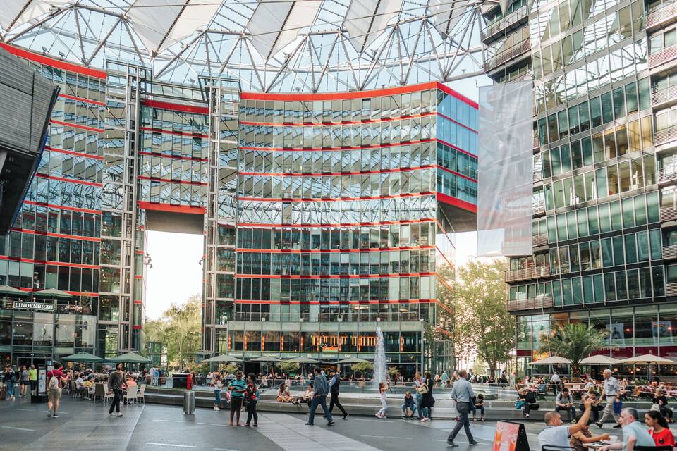 Een modern stadsplein met glazen gebouwen, terrasjes en een fontein.