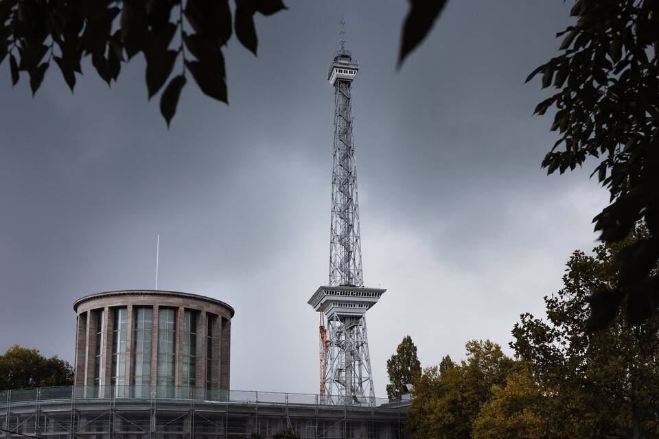 A tall tower with a spire, surrounded by trees and a building with a circular base.