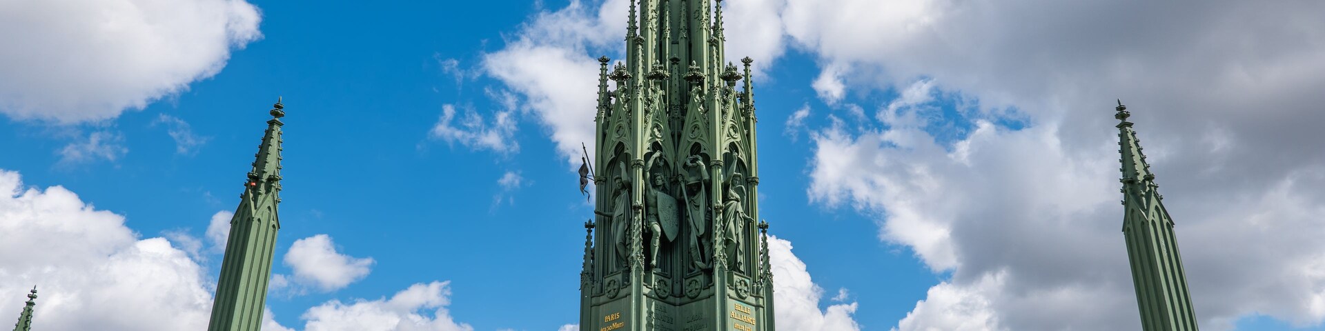 Uma torre em estilo gótico com pináculos e estátuas elaboradas, cercada por uma grade verde e degraus de pedra.