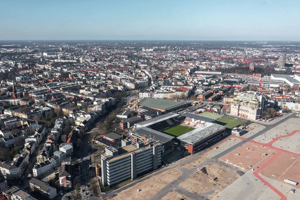 An aerial view of a city with a large stadium surrounded by dense urban buildings and a clear sky.