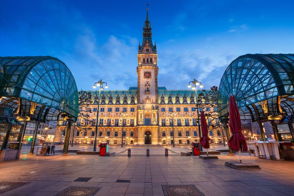 A historic building with a prominent clock tower, surrounded by a covered plaza with glass structures and street lamps.