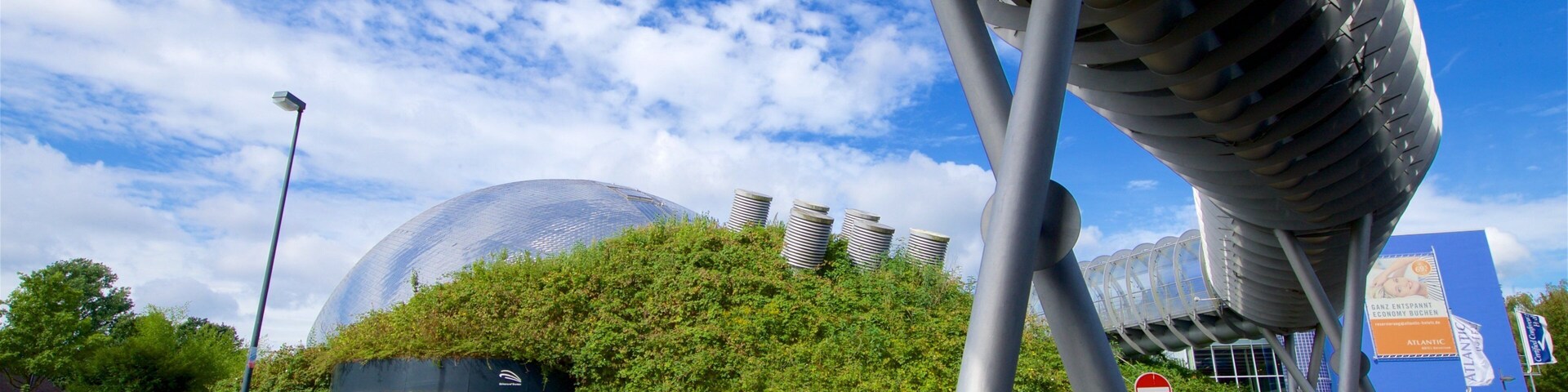 A modern building with a large dome and cylindrical structures, surrounded by greenery and a clear blue sky.