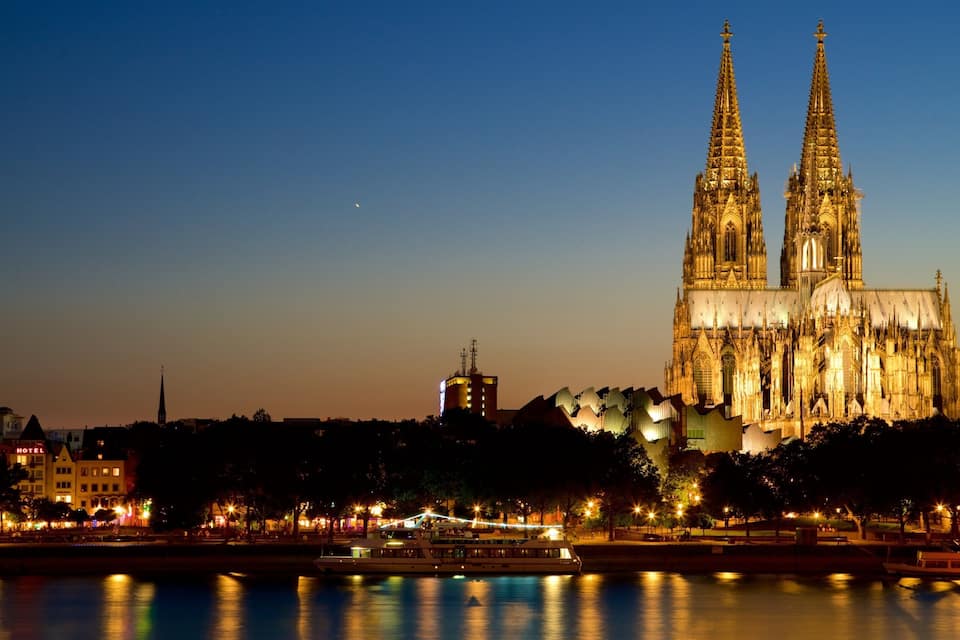 Une cathédrale avec deux hautes flèches, une rivière où naviguent des bateaux et un paysage urbain avec des bâtiments et des arbres.