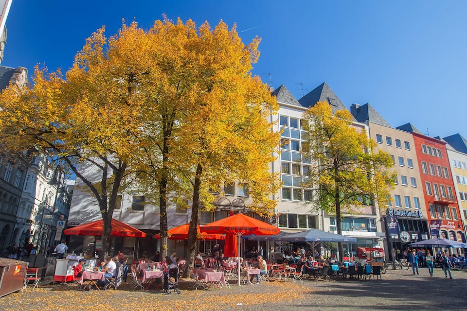 Une place pavée, avec des cafés en plein air, des arbres aux couleurs automnales et des bâtiments historiques.
