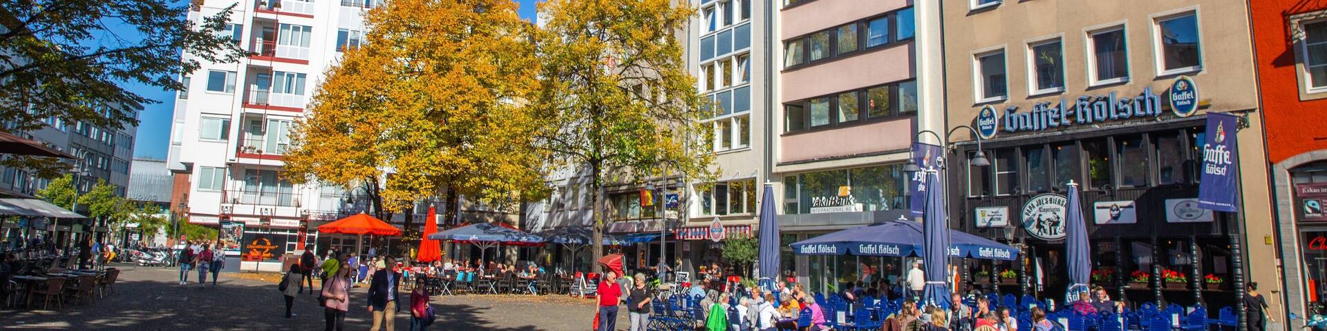 A bustling square with outdoor cafes, people walking, and autumn-colored trees.