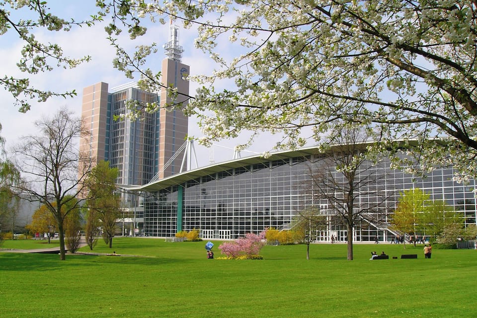 A modern building with a glass facade and a curved roof, surrounded by a green park with trees and a clear sky.