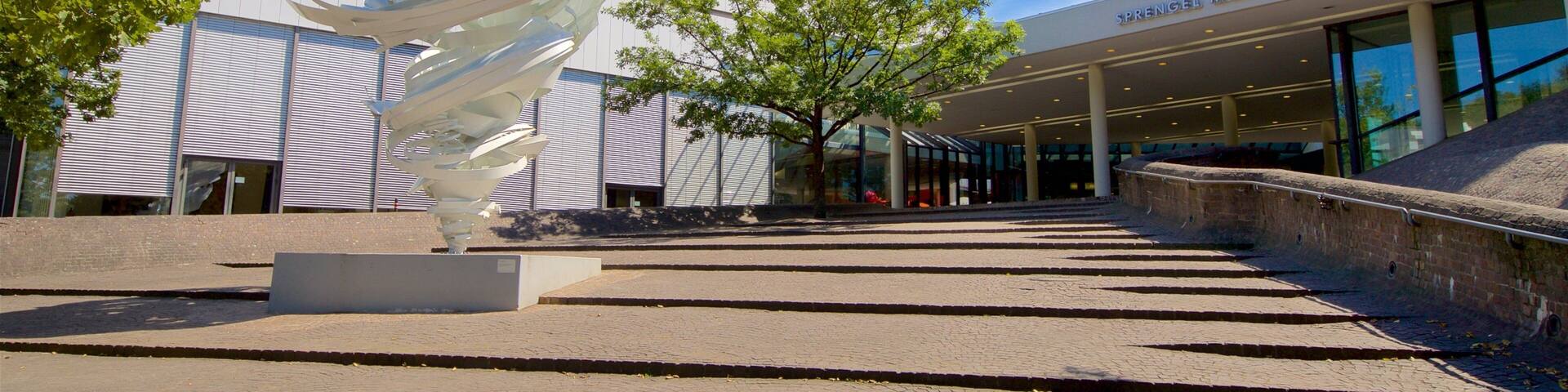 A modern building with a spiral sculpture in front, a tree, and a clear blue sky.