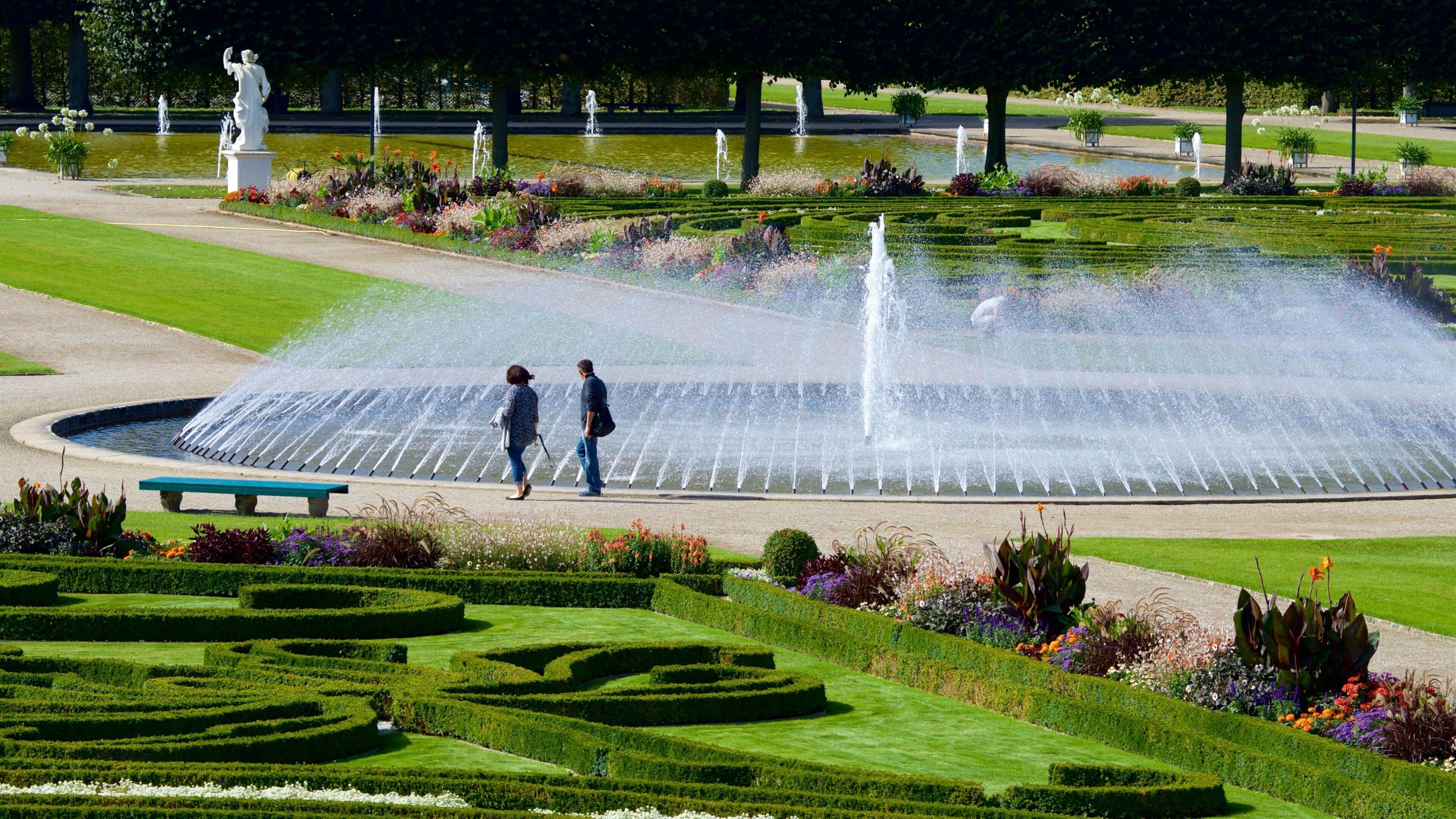 A large fountain with water jets, surrounded by manicured gardens and statues.