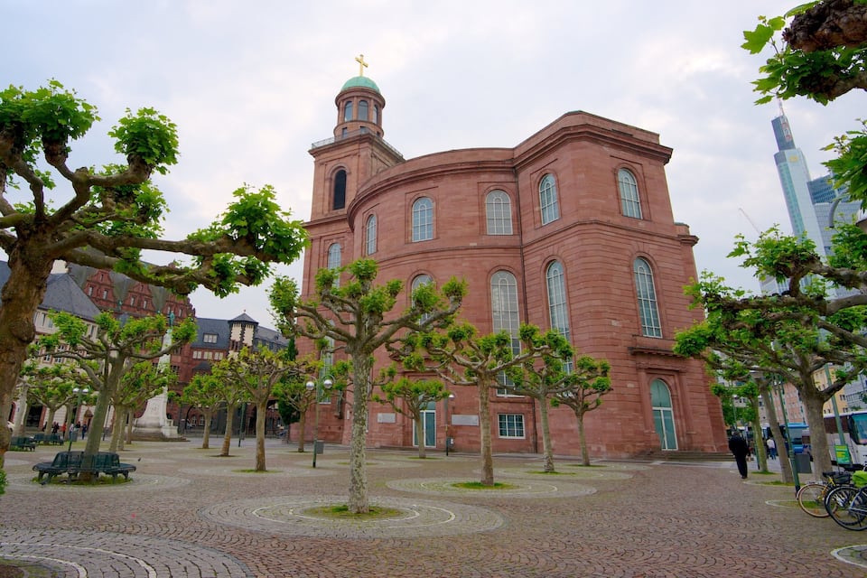A historic red brick building with a dome and cross, surrounded by trees and cobblestone paths.