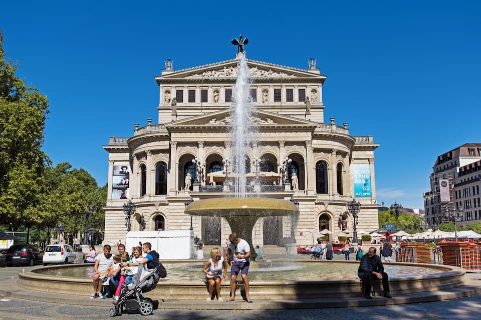 Edifici històric amb una font central, envoltat de gent i arbres.