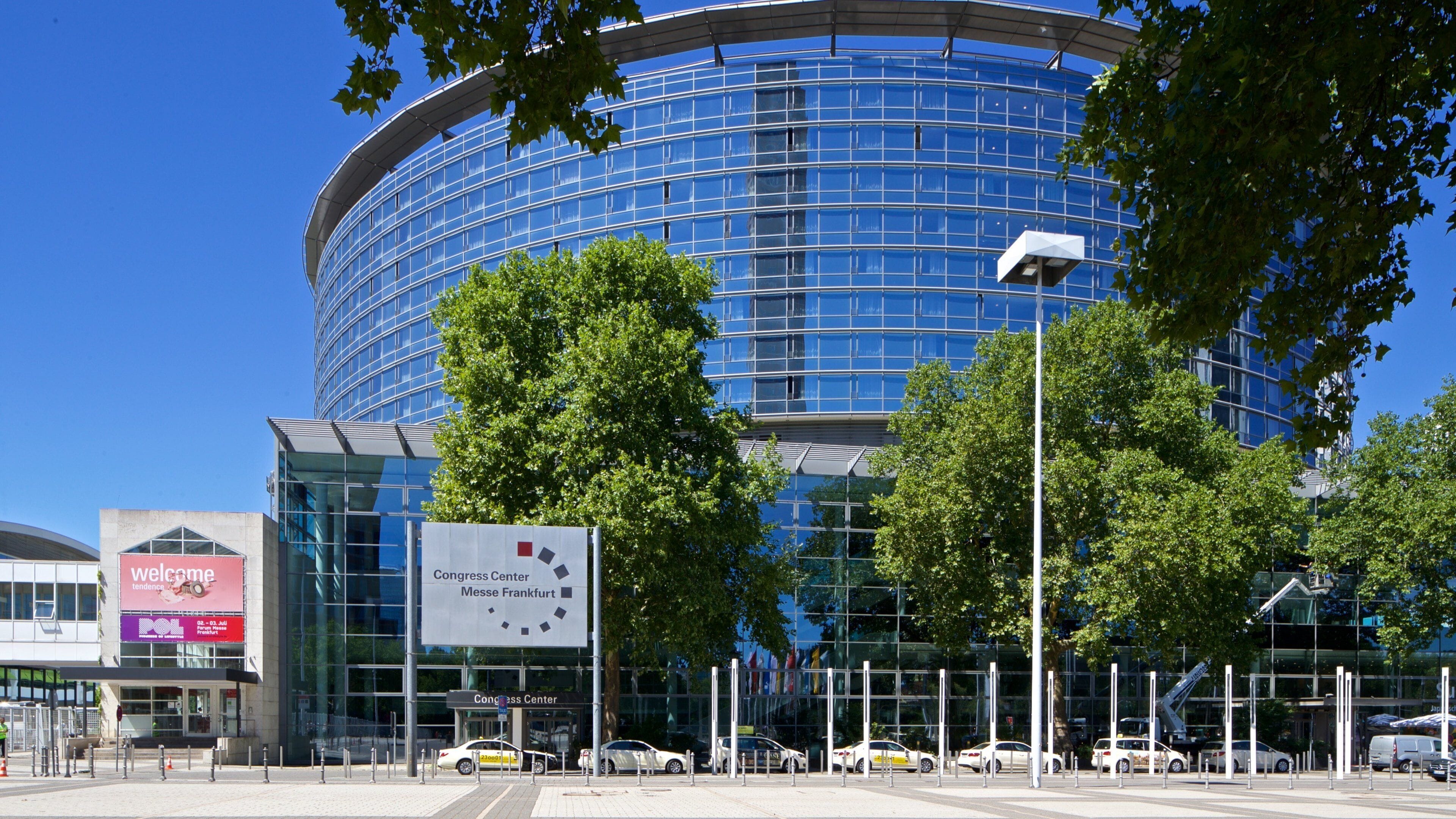 A modern glass building with a curved facade, surrounded by trees and a parking area with cars.