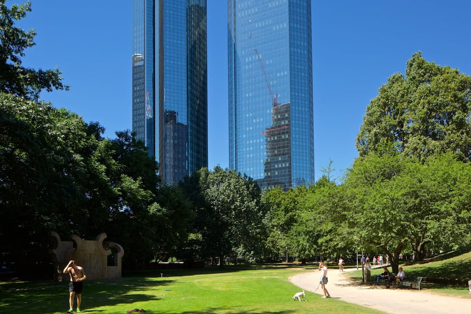 A park with green grass, trees, and a few people walking. In the background, there are tall buildings with glass windows.