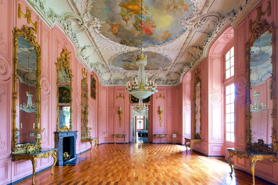 Une vaste pièce aux murs rose décorés, dotée d’un parquet en bois et d’un plafond orné d’une fresque détaillée.