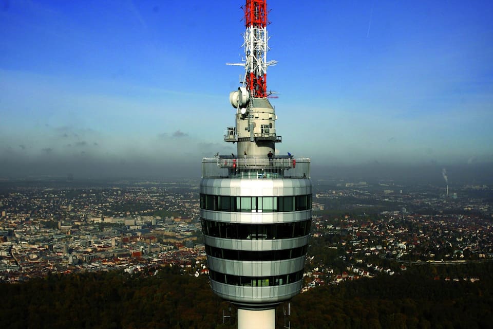 A tall tower with a red antenna, surrounded by a cityscape and forested area.