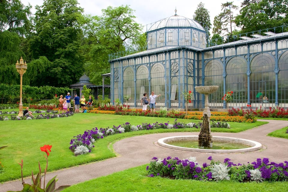 Een serre met een koepel, omgeven door een verzorgde tuin met diverse bloemen en een centrale fontein.