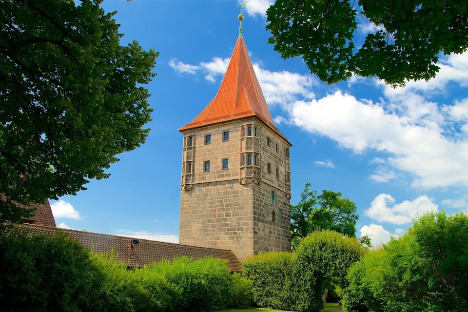 Uma torre histórica com telhado vermelho e paredes de pedra, rodeada por árvores verdes exuberantes e um céu azul límpido.