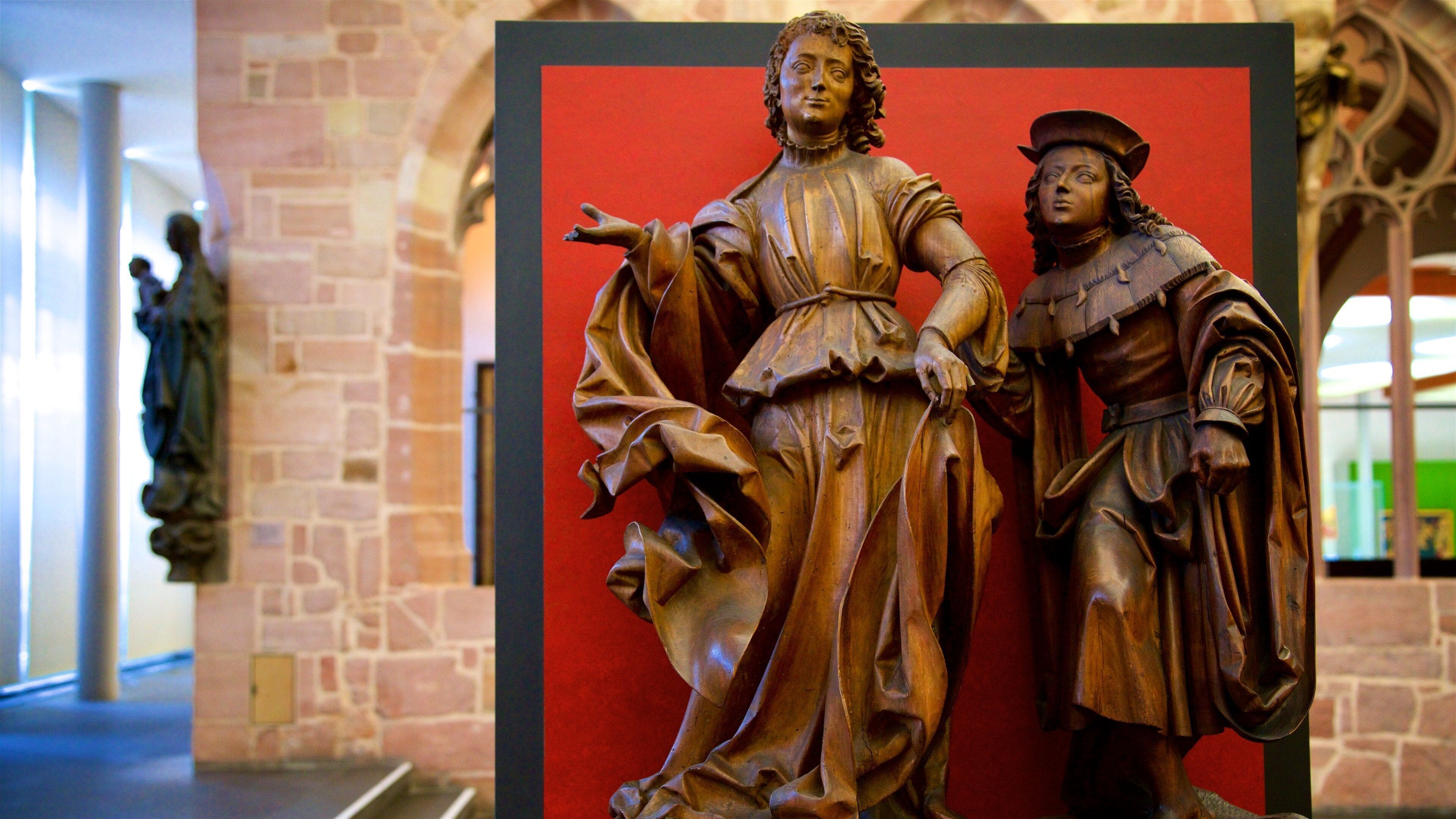 Une sculpture en bois représentant un homme et une femme vêtus de costumes d’époque, placée devant un fond rouge dans un bâtiment en pierre.