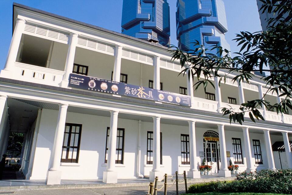 A white colonial-style building with a signboard in a foreign language, surrounded by modern skyscrapers.