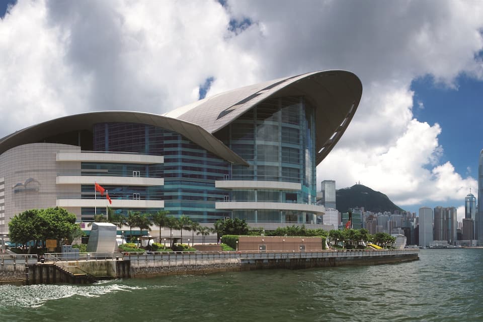 A modern architectural building with a curved design, situated by a waterfront with a cityscape in the background.